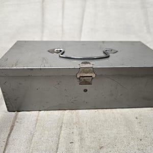 May include: A rectangular, gray metal toolbox with a handle and a latch. The toolbox has a slightly worn appearance, suggesting it has been used. The handle is a curved metal bar, and the latch is a rectangular piece of metal.