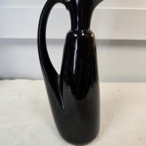Könnte beinhalten: Eine glänzend schwarze Kanne mit einem gebogenen Griff und einem Ausguss. Die Kanne hat einen hohen, schlanken Körper und eine glatte, reflektierende Oberfläche. Der Griff ist elegant geschwungen und der Ausguss ist für einfaches Ausgießen konzipiert.