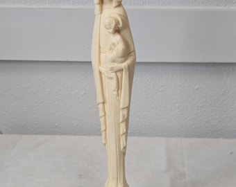 May include: Ivory-colored statue of the Virgin Mary holding a child, with a halo above her head. The statue has a black base and is made of a smooth material. The Virgin Mary is wearing a draped robe.