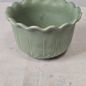 May include: A light green ceramic bowl with a scalloped rim and vertical ribbed detailing. The bowl has a rounded shape and is likely used for decorative purposes or as a planter. The color is a muted, pastel green.