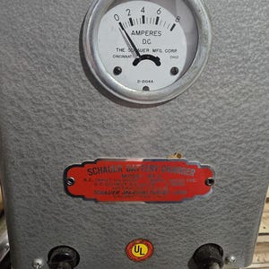 以下が含まれることがあります： ヴィンテージのグレーのバッテリー充電器のクローズアップ。 「AMPERES D.C.」と表示された丸いアナログメーターと、「SCHAUER BATTERY CHARGER」と書かれた赤と白のラベルが付いています。 充電器には2本の黒い電源コードが付いています。