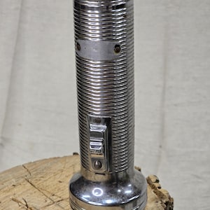 May include: A vintage, chrome-plated flashlight with a ribbed body and a rectangular switch. The flashlight has a circular base and a top with a small opening. It is displayed on a wooden surface.