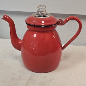 May include: A red enamel teapot with a clear glass lid and a curved handle. The teapot has a spout and a black rim around the lid. The teapot is a classic design.