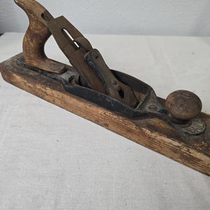 May include: A vintage wood plane with a wooden handle and knob. The plane has a metal blade and a dark metal frame. The wood shows signs of wear and age. The plane is a carpentry tool.