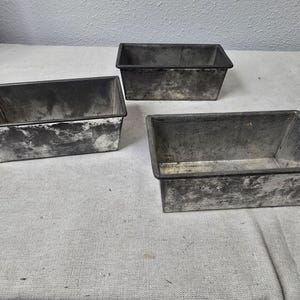 May include: Three rectangular metal loaf pans with a distressed, aged appearance. The pans are various sizes and sit on a light-colored surface. The pans have a dark rim and show signs of wear.