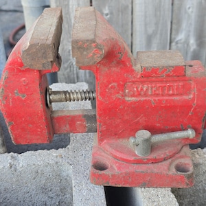 May include: A red Wilton bench vise, showing signs of wear. The vise has a screw mechanism and a handle for tightening. The jaws are open, ready to hold a workpiece. The vise is sitting on a concrete block.