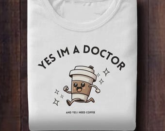 May include: White t-shirt with the text "YES IM A DOCTOR" above a cartoon coffee cup running with a smiling face. Below the coffee cup is the text "AND YES I NEED COFFEE". The design includes star accents.
