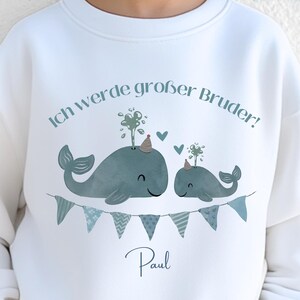 May include: White sweatshirt with a whale graphic and the text "Ich werde gro&szlig;er Bruder!" and the name "Paul". The graphic includes two whales, bunting, and hearts. The text "viele Farben" is also visible.
