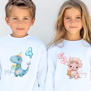 Puede incluir: Dos sudaderas blancas con dise&ntilde;os de dinosaurios. Una presenta un dinosaurio azul con un globo con el n&uacute;mero 4 y el nombre "Matteo". La otra tiene un dinosaurio rosa con un globo con el n&uacute;mero 5 y una magdalena, con el nombre "Romy".