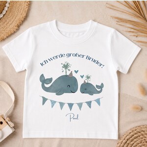 May include: A white short-sleeved t-shirt featuring a graphic of two blue whales wearing party hats, with the German text Ich werde gro&szlig;er Bruder! above them, a blue bunting banner below, and the name Paul printed at the bottom of the design.