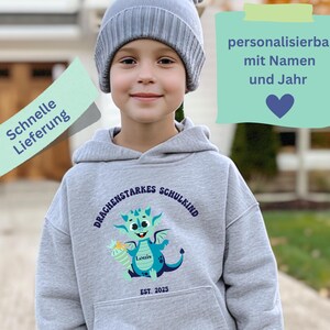 Pode incluir: Uma criança vestindo um moletom cinza com um desenho de dragão e o texto "DRACHENSTARKES SCHULKIND" e "EST. 2025". O moletom também tem o nome "Louis". A criança também está usando um gorro cinza.