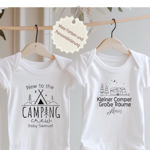 Könnte beinhalten: Zwei weiße Baby-Bodys hängen an Holzbügeln. Einer zeigt ein Camping-Design mit dem Text "New to the CAMPING crew Baby Samuel". Der andere hat ein Wohnmobil-Design mit deutschem Text.