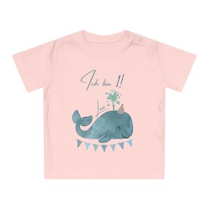 May include: A light pink baby t-shirt with a watercolor illustration of a blue whale wearing a party hat. The text "Ich bin 1! Levi" is written above the whale. A banner of blue and white triangles is below the whale.