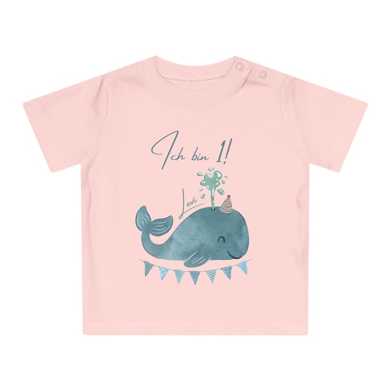 May include: A light pink baby t-shirt with a watercolor illustration of a blue whale wearing a party hat. The text "Ich bin 1! Levi" is written above the whale. A banner of blue and white triangles is below the whale.