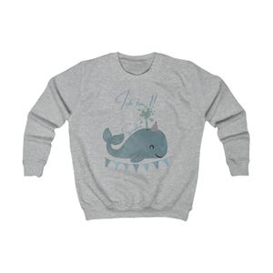May include: A light gray sweatshirt featuring a blue whale illustration with a party hat and water spout. The text "Ich bin 1!" is above the whale, and a banner with blue flags is below. A cute design for a child's birthday.