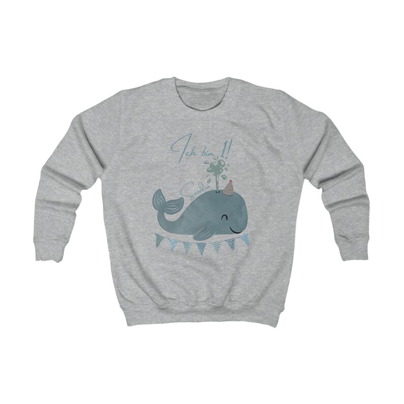 May include: A light gray sweatshirt featuring a blue whale illustration with a party hat and water spout. The text "Ich bin 1!" is above the whale, and a banner with blue flags is below. A cute design for a child's birthday.