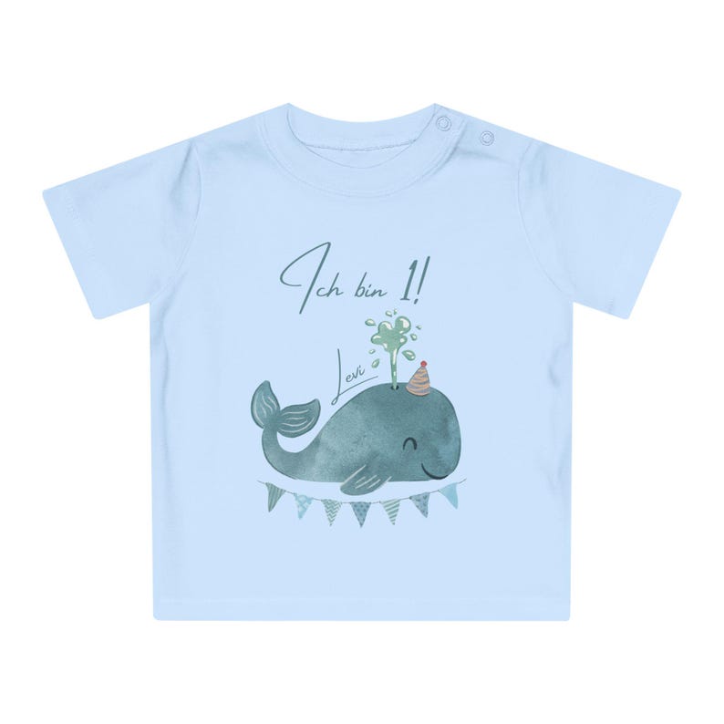 May include: Light blue baby t-shirt with a watercolor illustration of a whale wearing a party hat. The text "Ich bin 1! Levi" is written above the whale. A banner of triangular flags is below the whale.