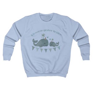 May include: A light blue sweatshirt featuring a graphic of two whales wearing party hats and a banner. The text "Ich werde gro&szlig;er Bruder!" is above the whales. The whales are light green with a light green banner.