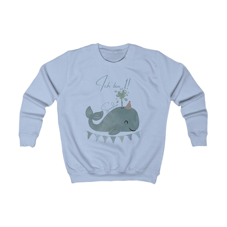 May include: Light blue sweatshirt featuring a cartoon whale design. The whale is gray-green and wears a party hat. The text "Ich bin 1!" and the name "Sandie" are above the whale. A banner with triangular flags is below the whale.