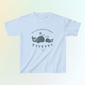 May include: A light blue t-shirt featuring a graphic of two grey whales wearing party hats, with the German text "Ich werde gro&szlig;er Bruder!" above them, a small heart, and a string of blue and grey triangular bunting flags below, with the name "Paul".