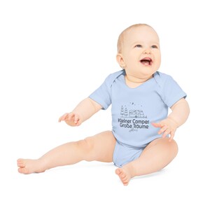 May include: A light blue baby onesie with short sleeves. The onesie features a black line drawing of a camper, trees, and text that reads "Kleiner Camper Gro&szlig;e Tr&auml;ume". The baby is sitting and smiling.