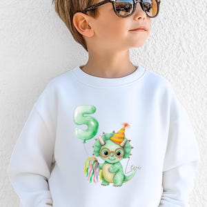 Puede incluir: Sudadera blanca con un dinosaurio verde con gafas y un gorro de fiesta, sosteniendo una serpentina colorida y un globo verde con el n&uacute;mero 5. El nombre "Levi" est&aacute; escrito en cursiva. Para ni&ntilde;os.
