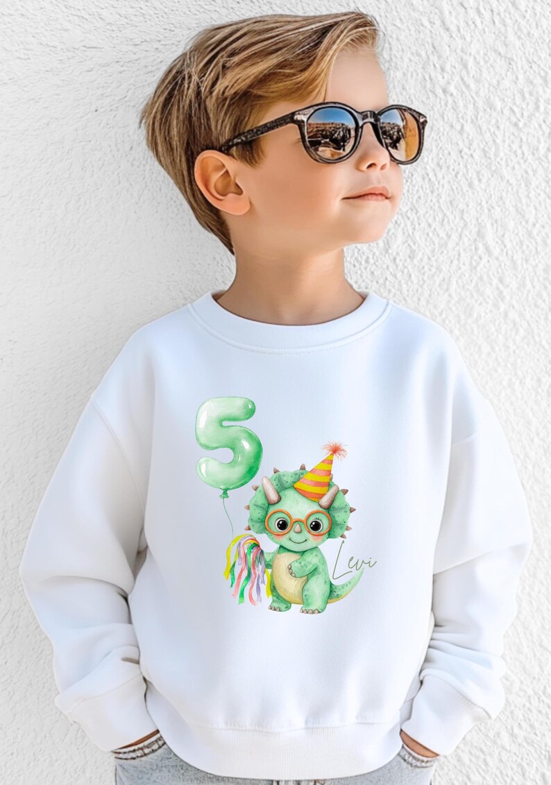 Puede incluir: Sudadera blanca con un dinosaurio verde con gafas y un gorro de fiesta, sosteniendo una serpentina colorida y un globo verde con el n&uacute;mero 5. El nombre "Levi" est&aacute; escrito en cursiva. Para ni&ntilde;os.