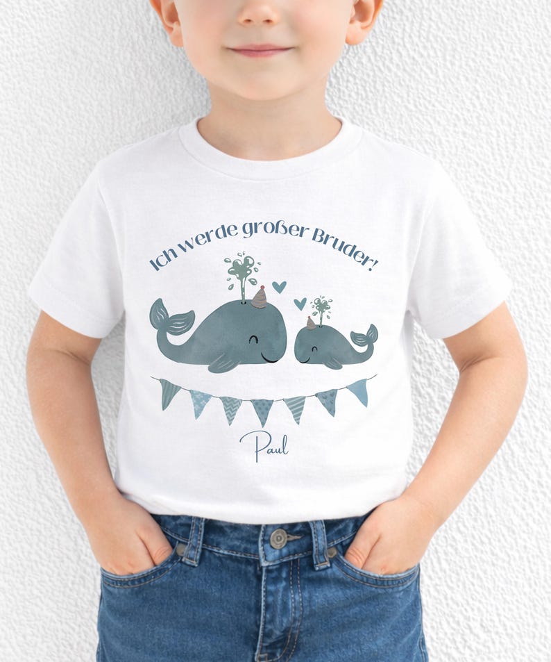 May include: A white short-sleeved t-shirt featuring a graphic of two blue whales wearing party hats, with water spouts, hearts, and a bunting banner. The text reads "Ich werde gro&szlig;er Bruder!" and the name "Paul" is printed below the banner.