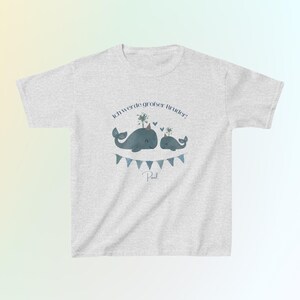 May include: A light gray short-sleeve t-shirt featuring a graphic of two blue whales wearing party hats, with a blue bunting banner below them. The text "Ich werde gro&szlig;er Bruder!" is printed above the whales, and the name "Paul" is below.