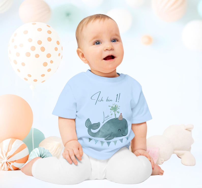 May include: A baby wearing a light blue t-shirt with a whale graphic and the text "Ich bin 1!". The whale has a party hat and a water spout with a shamrock. The baby is sitting in front of balloons and paper decorations.