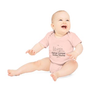May include: A light pink baby bodysuit with a black graphic of a camper, trees, and text that reads "Kleiner Camper Gro&szlig;e Tr&auml;ume". The baby is sitting and smiling.