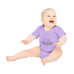 May include: A light purple baby bodysuit with short sleeves. The bodysuit features a black graphic of a camper, trees, and text that reads "Kleiner Camper Gro&szlig;e Tr&auml;ume". The baby is sitting and smiling.