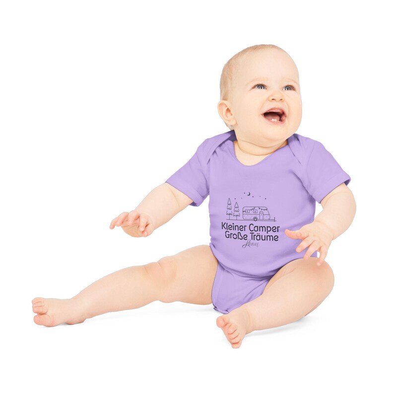 May include: A light purple baby bodysuit with short sleeves. The bodysuit features a black graphic of a camper, trees, and text that reads "Kleiner Camper Gro&szlig;e Tr&auml;ume". The baby is sitting and smiling.