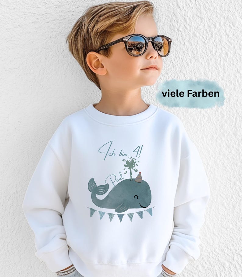 May include: White sweatshirt with a whale graphic, a party hat, and the text "Ich bin 4!" and "Paul". The whale is blue-green with a banner of small flags below. The text "viele Farben" is in a blue rectangle.