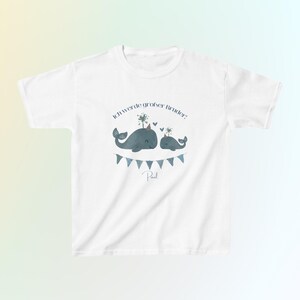 May include: A white short-sleeved t-shirt featuring a graphic of two blue whales, one larger than the other, wearing party hats. Above the whales is the text 'Ich werde gro&szlig;er Bruder!' and below them is a blue bunting banner with the name 'Paul'.