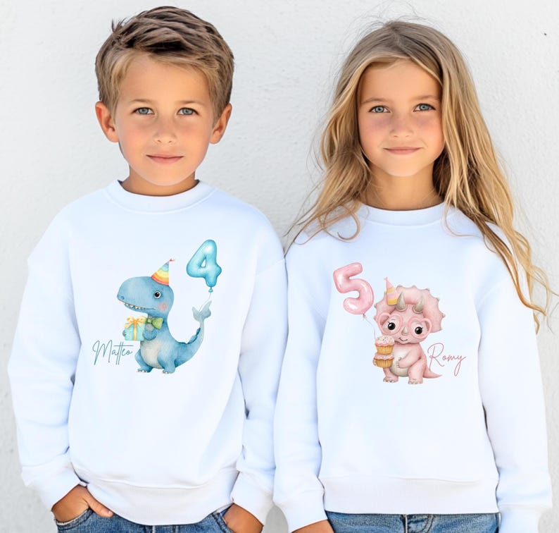 Puede incluir: Dos sudaderas blancas con dise&ntilde;os de dinosaurios. Una presenta un dinosaurio azul con un globo con el n&uacute;mero 4 y el nombre "Matteo". La otra tiene un dinosaurio rosa con un globo con el n&uacute;mero 5 y una magdalena, con el nombre "Romy".