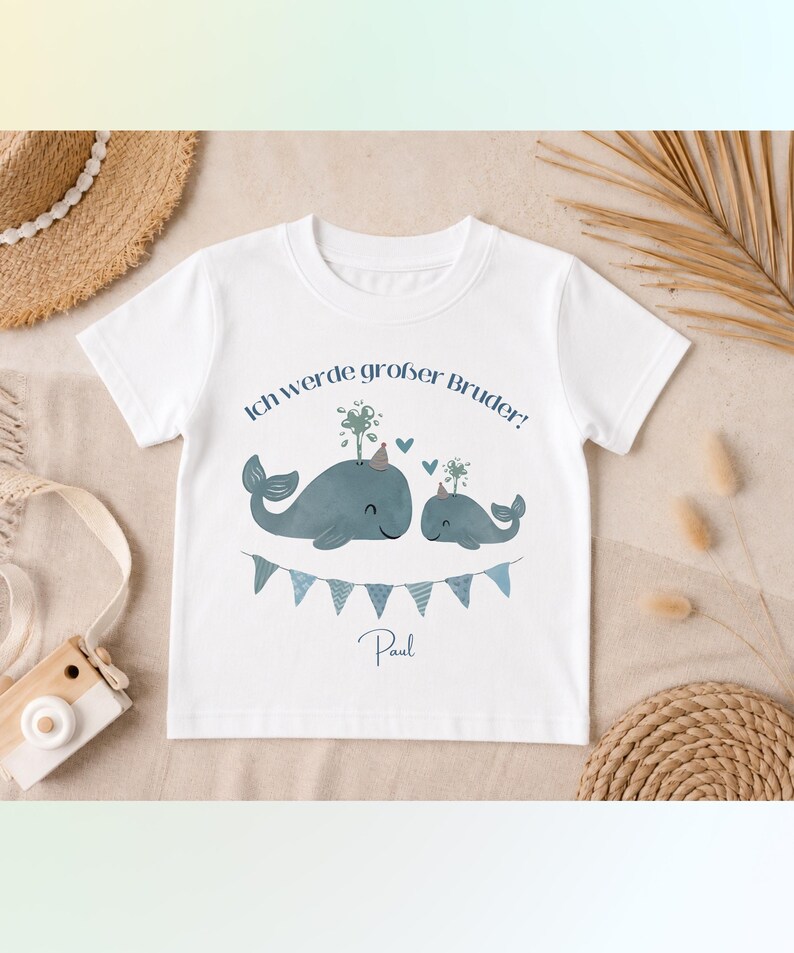 May include: A white short-sleeved t-shirt featuring a graphic of two blue whales wearing party hats, with the German text Ich werde gro&szlig;er Bruder! above them, a blue bunting banner below, and the name Paul printed at the bottom of the design.