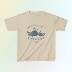May include: A beige short-sleeve t-shirt featuring a graphic of two blue whales with party hats, a string of blue triangular bunting, and the German text Ich werde gro&szlig;er Bruder! with the name Paul printed in a dark blue script font.