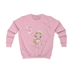 Puede incluir: Sudadera rosa claro con un dinosaurio de dibujos animados sosteniendo un cono de helado y usando gafas. Un globo rosa con el n&uacute;mero 5 est&aacute; a la izquierda. El nombre "Romy" est&aacute; escrito en cursiva a la derecha.