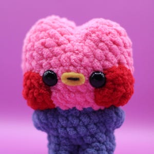 Puede incluir: Una muñeca de ganchillo hecha a mano con una cabeza rosa en forma de corazón, mejillas rojas, ojos negros y un pico amarillo. La muñeca tiene un cuerpo azul y está sobre un fondo morado. Este peluche es un artículo lindo y tierno.