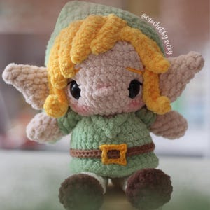 Puede incluir: Muñeca de ganchillo hecha a mano de un personaje con túnica verde, cinturón marrón y gorro puntiagudo. La muñeca tiene pelo rubio, ojos negros grandes y pequeñas alas beige. El texto "@crochetbyvicky" es visible.