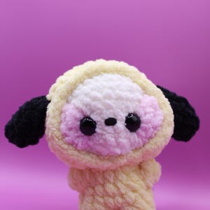 Puede incluir: Un peluche hecho de hilo suave, con una cabeza redonda con capucha de color amarillo pálido, una cara blanca y rosa claro, y dos orejas negras. El juguete tiene ojos negros pequeños y una nariz negra diminuta, con cuerpo amarillo pálido y brazos cortos.