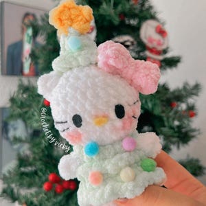 Puede incluir: Una figura blanca hecha a mano con un lazo rosa, ojos negros y un pico amarillo. La figura lleva un vestido verde claro adornado con pompones de colores. Un pequeño sombrero naranja y verde está en la parte superior de su cabeza. Un árbol de Navidad en el fondo.