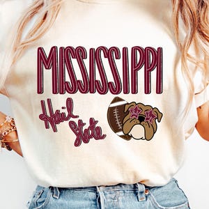 May include: A cream-colored t-shirt featuring the text MISSISSIPPI in maroon, the phrase Hail State in maroon script, and a graphic of a brown bulldog wearing star-shaped glasses next to a brown and white football with white laces.