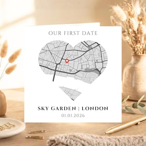 May include: A white card with the text "OUR FIRST DATE" above a heart-shaped map of London, with a red marker. Below, it reads "SKY GARDEN | LONDON" and the date "01.01.2026". The card is on a wooden surface.