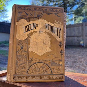 May include: A hardcover book titled Museum of Antiquity with a tan cover featuring black and gold illustrations of ancient structures, a central circular emblem depicting three figures, and a decorative border with geometric patterns at the base.