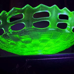 May include: A glowing green glass basket with a woven pattern on the base and oval cutouts around the rim. The basket has a scalloped edge and is illuminated, creating a vibrant display. This decorative piece is ideal for displaying fruit or other items.