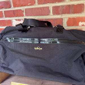 May include: A black Kipling duffel bag featuring a horizontal glossy black trim across the front, a top zipper closure, two black fabric handles, and a small yellow logo embroidered on the center. A small white plush monkey keychain hangs from the side.