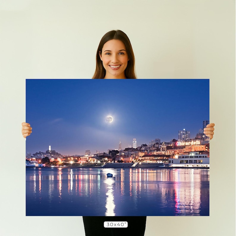 A woman is holding up a large framed print of a nighttime cityscape with a full moon and boats on the water.