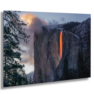 May include: A landscape photograph of a waterfall illuminated by the setting sun, creating an orange glow. The waterfall cascades down a large rock formation, surrounded by trees and a cloudy sky. The image captures a natural phenomenon.